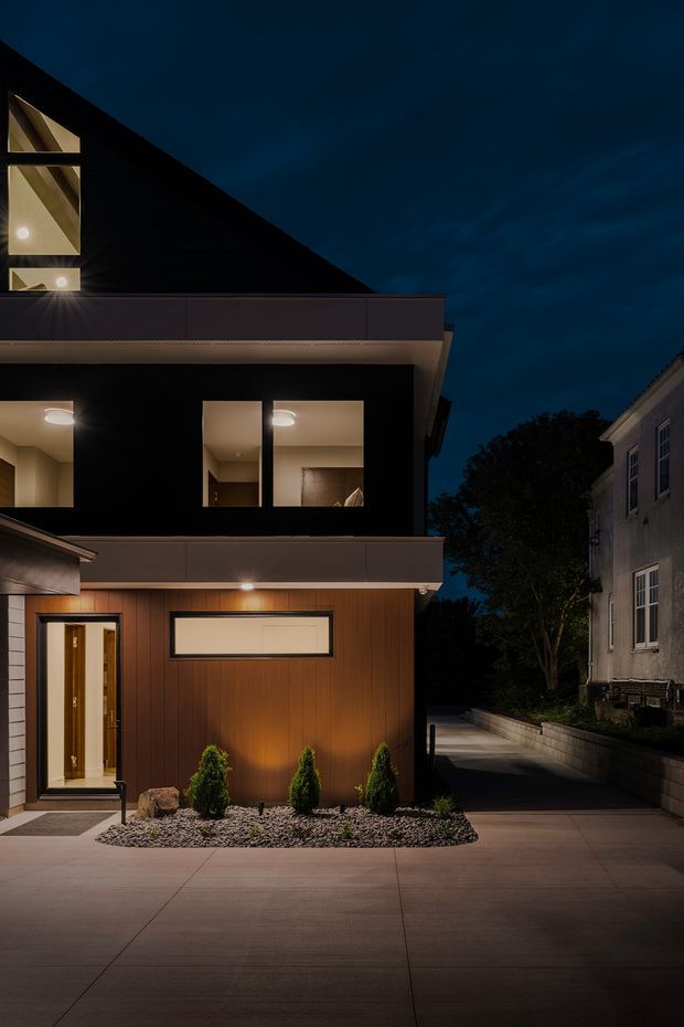 Night-time exterior view of a contemporary multi-level residence. The dark, sharply pitched roof and upper-level glazing contrast with a warm vertical timber-clad lower facade, illuminated entry, projecting flat-roofed balcony or canopy, and a broad concrete driveway. A neighbouring traditional house is visible to the right, with low planting and pebble landscaping along the frontage.
