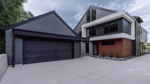 Contemporary multi-level residential exterior with a dark horizontal-clad gabled garage wing adjoining a taller main volume. The facade combines charcoal cladding, white rendered or framed bands, extensive dark-framed glazing, and a warm timber-toned entry wall. A broad pale concrete driveway and minimal gravel planting complete the clean, modern composition.