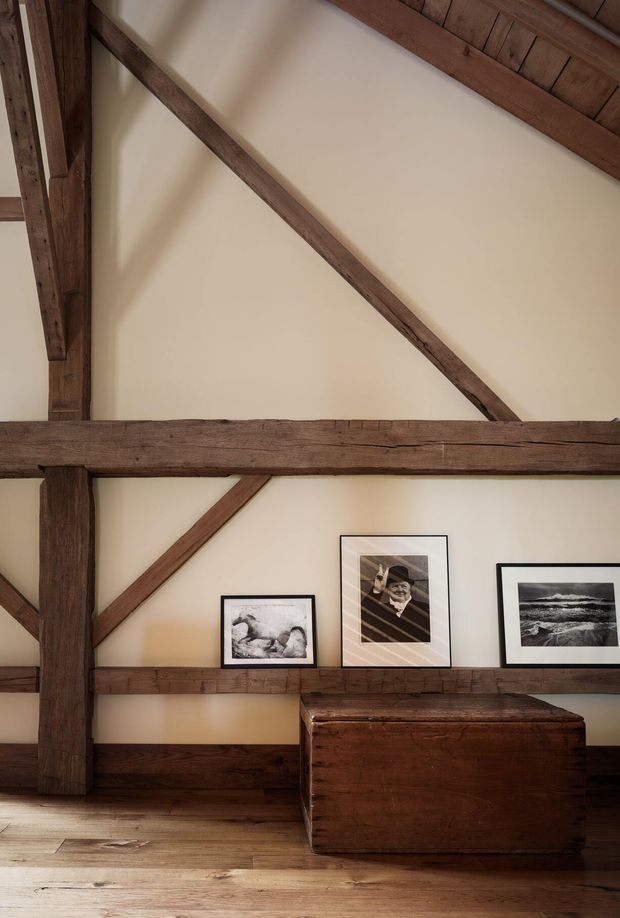 Interior view of a rustic timber-framed room with exposed dark wood beams, cream plaster infill walls, and a warm timber floor. A long built-in timber ledge displays framed black-and-white artwork, with a substantial dark wood storage chest or bench positioned below. The composition highlights the dramatic structural framing and warm, historic character.