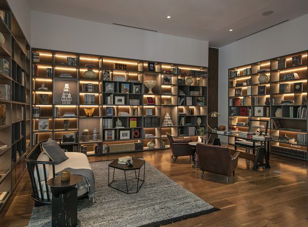 A sophisticated living room or study area dominated by full-height built-in shelving and cabinetry with warm integrated LED lighting. The shelves display books, artwork, ceramics, and decorative objects. Leather lounge chairs, a sofa, side tables, a desk, and a large textured rug create a refined library-like seating area. The room has dark timber shelving, warm timber flooring, neutral walls, recessed ceiling lights, and a calm, moody atmosphere.
