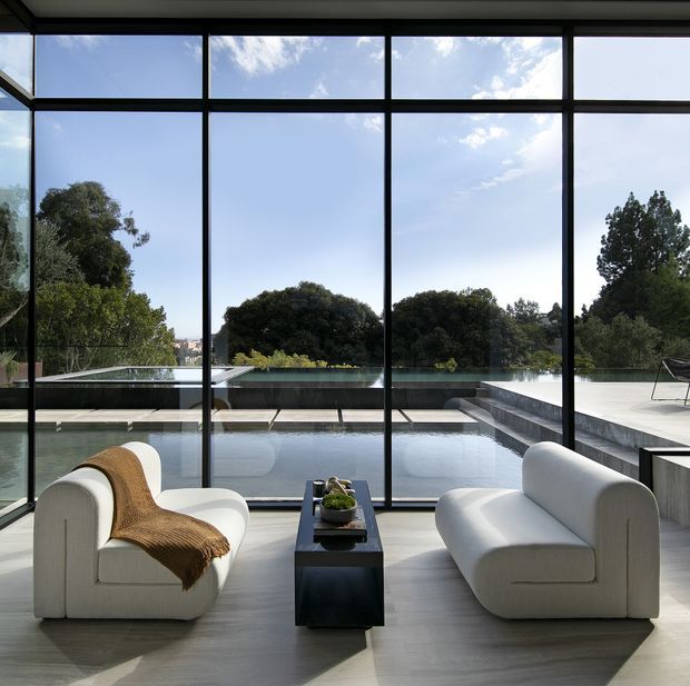 A refined living room with two sculptural cream lounge chairs and a narrow dark coffee table, arranged in front of expansive black-framed floor-to-ceiling glazing. The glazing overlooks a sleek outdoor swimming pool and terrace, with mature trees beyond. The interior has a restrained, contemporary composition with pale flooring, strong horizontal and vertical window divisions, and abundant natural light.