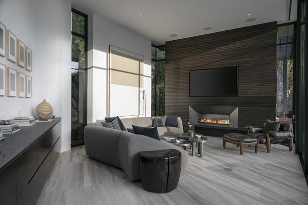 A refined contemporary living room with a large sectional sofa arranged around a linear fireplace and wall-mounted television. The room features floor-to-ceiling glazing, a dark horizontal timber feature wall, pale grey wood-look flooring, a minimalist black built-in sideboard, and sculptural lounge furniture. Natural light creates a bright yet sophisticated atmosphere.