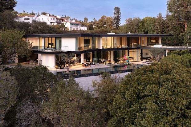 Exterior view of a large contemporary two-storey residence set among mature trees on a sloped, landscaped site. The house has a long, low flat roof, extensive floor-to-ceiling glazing, dark slim framing, white rendered wall volumes, balconies, and a partially visible swimming pool and terrace at the lower level. A neighbouring hillside residence is visible in the background.