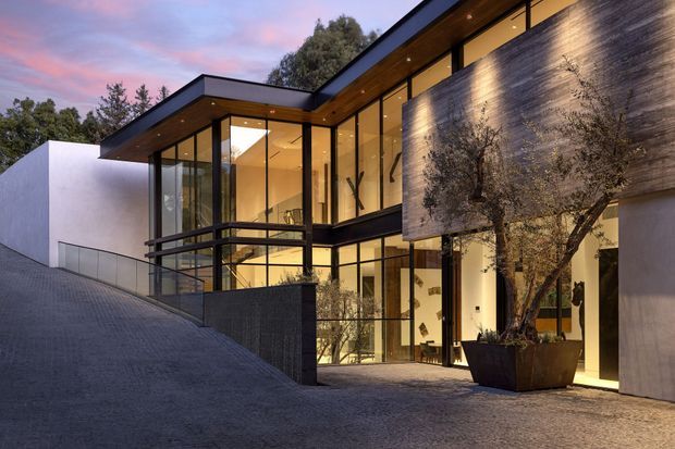 Contemporary multi-level residential facade at dusk, set on a sloped driveway. The building combines extensive floor-to-ceiling glazing with dark metal framing, flat roof planes, smooth white rendered walls, and a prominent textured concrete or stone feature wall. Warm interior lighting is visible through the glazing, with a small upper-level balcony and large planters at the entrance.