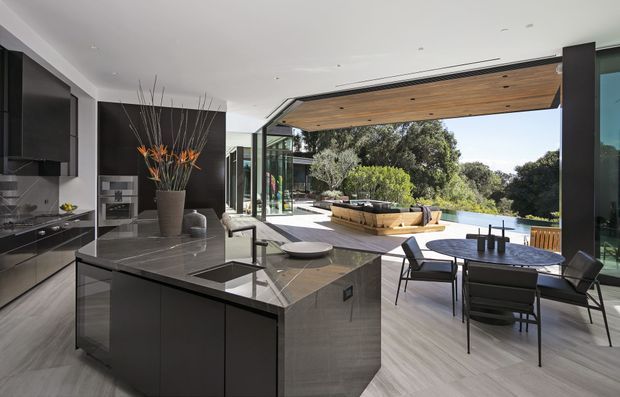 A high-end contemporary open-plan kitchen and dining area opening onto an outdoor terrace and poolside setting. The kitchen features a large dark island with an integrated sink and stone benchtop, dark fixed cabinetry, and a grey tiled or stone floor. Full-height retractable glazing creates a seamless connection to the terrace, which includes a timber-lined ceiling, dining table, built-in seating, and views toward a pool and landscaped trees.
