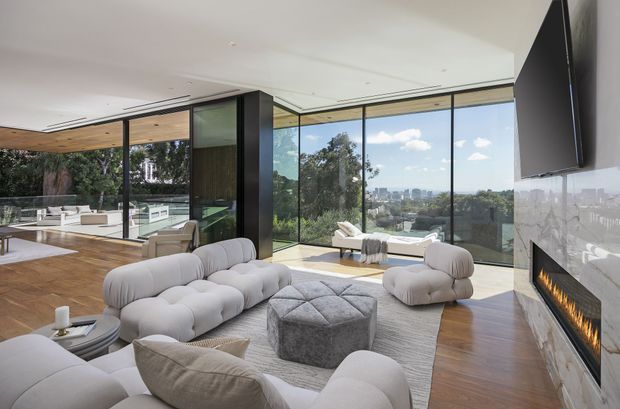 A spacious, high-end contemporary living room with panoramic floor-to-ceiling glazing overlooking a leafy urban landscape. The room features sculptural light-coloured sofas and armchairs arranged around a large tufted ottoman, warm timber flooring, a pale textured area rug, a sleek linear fireplace set into a polished pale stone wall, and a wall-mounted television. An open kitchen and dining area are visible beyond a dark-framed glazed partition, with timber ceiling detailing adding warmth.