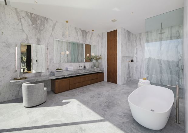 Luxurious contemporary bathroom featuring full-height grey-veined marble-look wall surfaces, matching large-format flooring, a long floating dark timber vanity with stone benchtop and twin basins, frameless mirrors, a freestanding white bathtub, and a frameless glass shower enclosure. A tall timber door and restrained brass-toned pendant and wall lighting add warmth to the bright, spacious room.