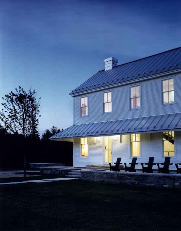 Exterior dusk view of a substantial two-storey white weatherboard house with a steep standing-seam metal gabled roof, chimney, evenly spaced double-hung windows, and a deep covered verandah. Dark outdoor lounge chairs sit on a stone terrace above a landscaped lawn, with trees surrounding the property.