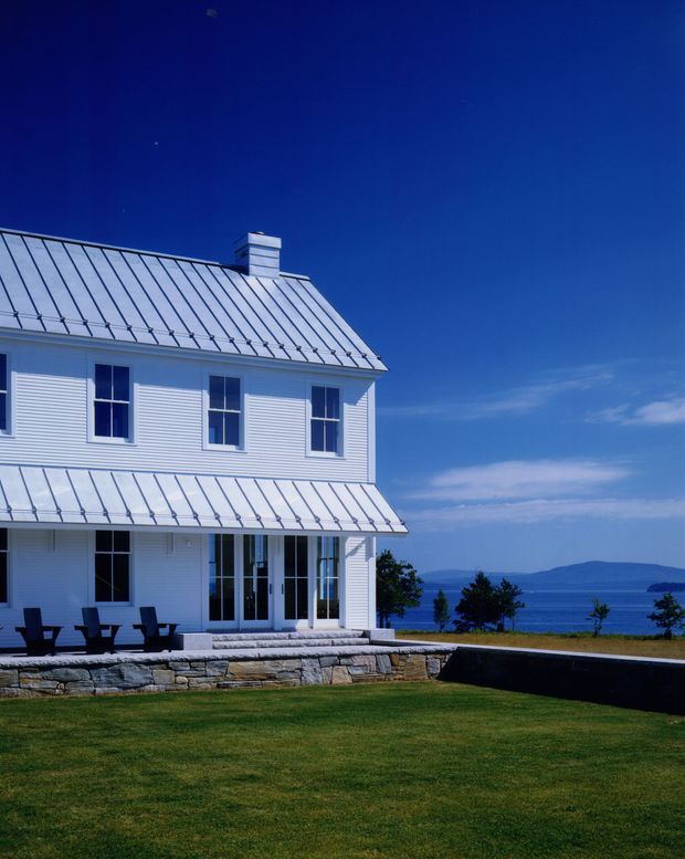 Exterior view of a two-storey white-clad house with a steep standing-seam metal roof, chimney, symmetrical windows, and a covered ground-floor terrace. The house sits on a low stone retaining wall overlooking a body of water and distant hills under a clear blue sky.