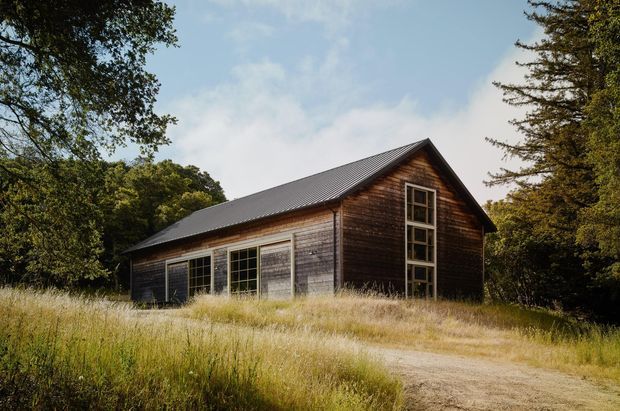 A rustic single-storey barn-like timber building set in a grassy rural landscape. The structure has dark weathered horizontal wood cladding, a steep gabled roof with dark metal roofing, large multi-pane glazed doors or windows along the side, and a tall vertical multi-pane window in the gable end. Mature trees surround the building.