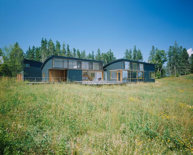 A contemporary single-storey house set within an open meadow and surrounded by trees. The building has a dark blue-grey horizontal-clad exterior, low curved and gabled roof forms, extensive floor-to-ceiling glazing, and warm timber-framed openings and outdoor decking. The broad frontage includes a sheltered central outdoor area and a glazed living wing.