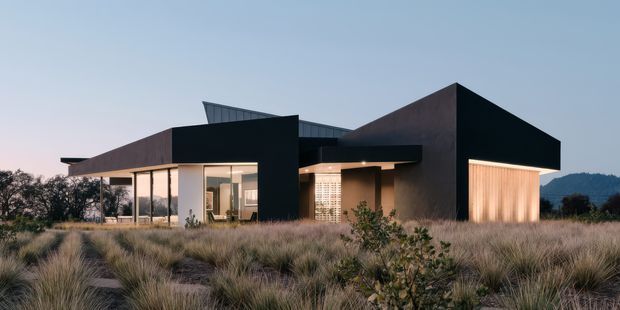 A low-slung contemporary house set within natural grasses, featuring bold geometric volumes, dark charcoal cladding, warm brown wall planes, extensive floor-to-ceiling glazing, and a projecting flat roof. Warm interior lighting glows through the glass and a sheltered entry, while the surrounding landscape and distant hills create a semi-rural setting.