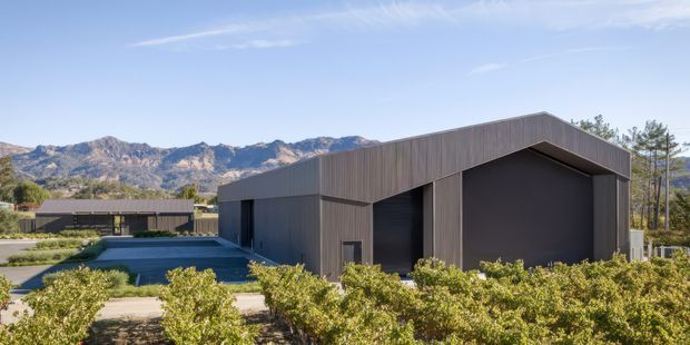 A contemporary single-storey building with a simple asymmetrical pitched roof and dark vertical cladding, set within landscaped grounds. Large recessed dark openings form the main facade, with a paved approach and low planting in the foreground. Rugged mountains are visible in the background.
