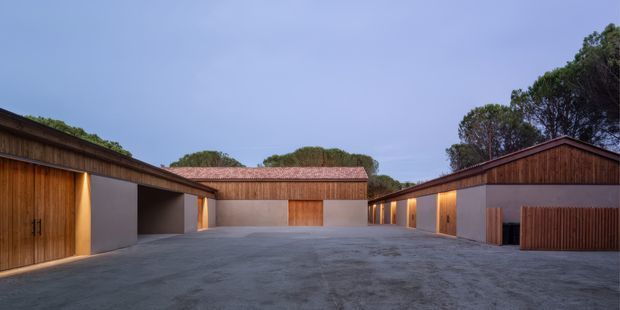 A dusk exterior view of a low-rise residential compound arranged around a broad paved courtyard. Several simple gabled buildings feature warm timber-clad sections, pale rendered walls, recessed garage or entry openings, and reddish tiled pitched roofs. Integrated lighting creates a warm glow against the cool blue sky, with mature trees visible behind the buildings.