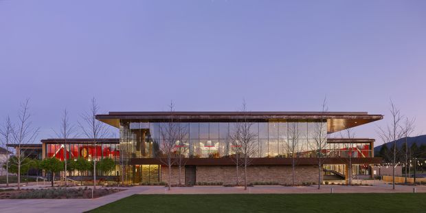 Wide exterior architectural photograph of a low-rise contemporary building at dusk. The structure features extensive floor-to-ceiling glazing, a broad projecting flat roof with thin horizontal profiles, warm timber-toned soffits, and a stone-clad central base. Sparse deciduous trees, paved paths, lawn, and surrounding landscape frame the building.
