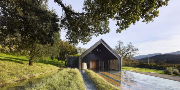A dark-clad, simple gable-roof building set within a landscaped rural or semi-rural site. The facade has a deep covered entry or open-sided porch with timber doors, while a broad reflective paved or water-like surface extends in front. Mature trees, ornamental grasses, and rolling hills frame the building.