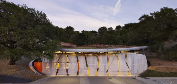 Exterior view of a distinctive sculptural, low-profile building set into a wooded hillside. The facade combines curved and angular structural elements, translucent or glazed wall panels, warm linear lighting, and a broad shallow roof with an organic, cave-like form. A paved forecourt occupies the foreground, while dense trees surround the structure.