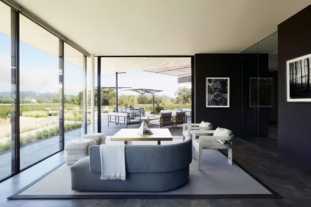 A refined contemporary living room opening onto a covered outdoor entertaining terrace through extensive floor-to-ceiling glazing. The room features a curved blue-grey sofa, pale lounge chairs, a low rectangular coffee table, a large light rug, dark feature walls and dark timber flooring. A landscaped rural view is visible beyond the terrace.