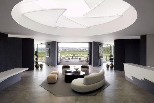 A spacious, sophisticated contemporary living room with dark feature walls, pale sculptural seating, and a low black circular coffee table arranged on a large geometric rug. A dramatic circular recessed ceiling feature with layered white geometric panels forms the main architectural focal point. Full-height glazing opens to an outdoor terrace and expansive landscape beyond, while built-in angular cabinetry or display units frame the room.