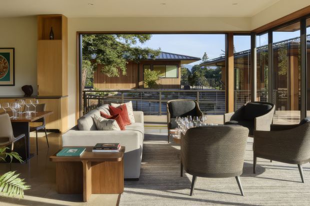 A warm, light-filled contemporary living room opening onto a balcony or terrace. The space features floor-to-ceiling timber-framed glazing, a pale sofa with red accent cushions, textured lounge chairs, a timber coffee table, built-in timber shelving, and a compact bar or dining area. Through the glazing is another timber-clad building and mature greenery.