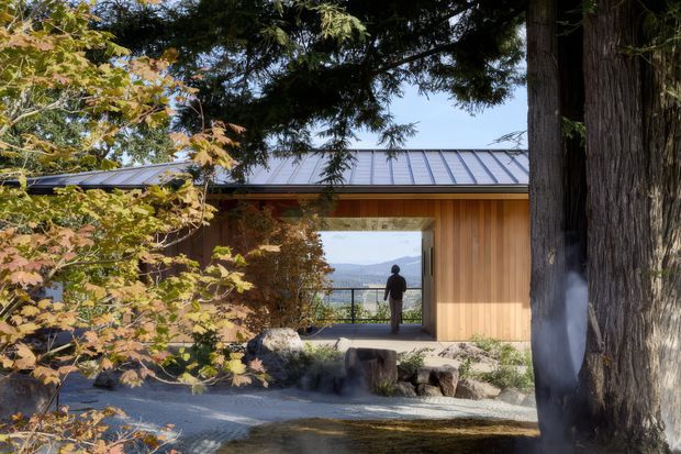 A low, single-storey timber-clad building set within a landscaped garden, viewed from outside through mature trees. The building has a dark standing-seam metal roof, warm vertical timber cladding, and a large glazed opening framing a distant mountain or water view. Natural rocks, gravel, logs, and established planting create a calm, Japanese-influenced garden setting.