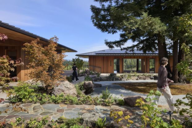 A landscaped courtyard connecting low-profile timber-clad buildings. The scene features broad overhanging roofs, extensive glazing, stone paving, gravel, large natural rocks, mature trees, and Japanese-inspired garden planting. People are walking through the courtyard, while the buildings appear to form a calm, contemporary retreat.
