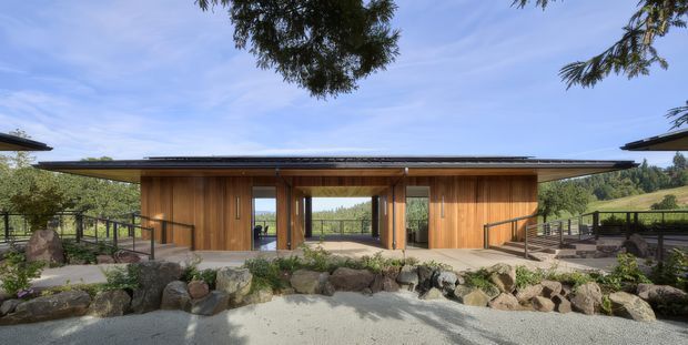 A low, single-storey pavilion-style building set within a landscaped rocky site. The facade features extensive warm vertical timber cladding, broad sheltered roof overhangs, large glazed openings, and a central open passage or courtyard connection. Dark metal railings and decking frame the structure, with planted slopes and trees beyond.