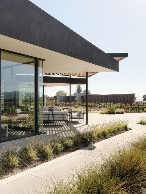 A contemporary single-storey residence with broad cantilevered roof planes, dark textured exterior fascia, extensive floor-to-ceiling glazing, and slim dark structural posts. The covered outdoor living area contains neutral lounge seating and opens onto a landscaped courtyard with ornamental grasses, paved walkways, and low planting. Strong horizontal lines, deep overhangs, and a bright, open indoor-outdoor connection define the scene.
