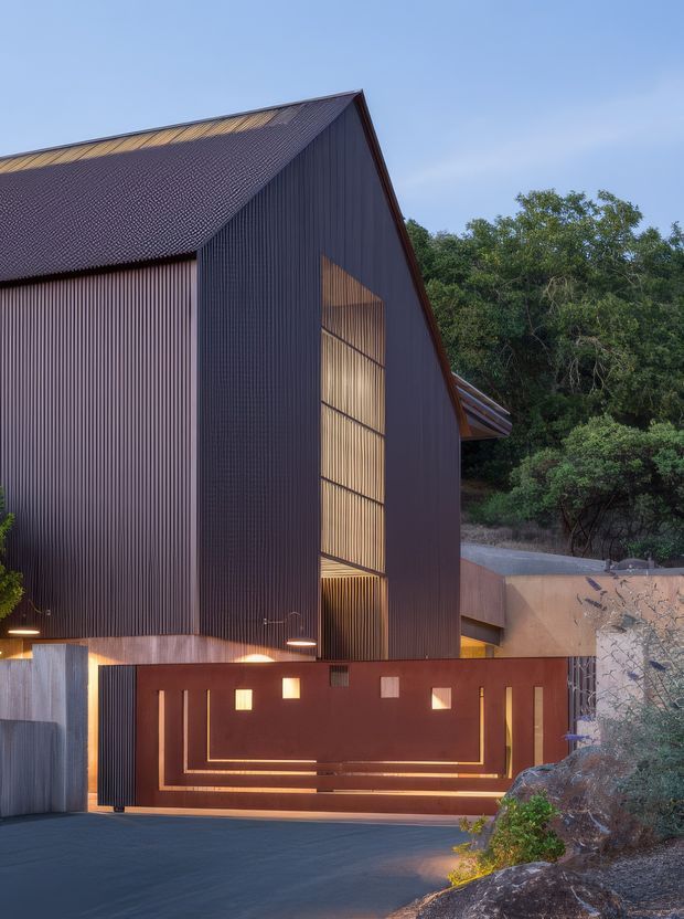 Exterior architectural photograph of a contemporary, barn-like building at dusk. The structure has a steep gabled roof, dark vertically ribbed metal cladding, a textured dark roof, and a tall warm-lit glazed or translucent opening. A rust-toned metal driveway gate with geometric illuminated details fronts the building, with landscaped bush vegetation and a paved driveway surrounding it.