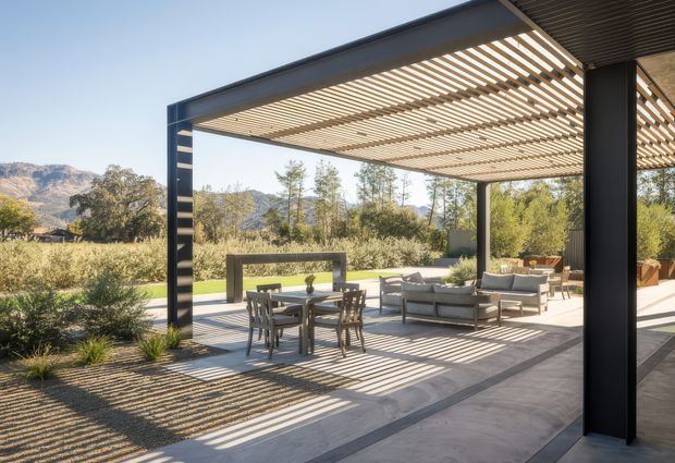 A spacious contemporary outdoor entertaining area beneath a large black steel pergola with closely spaced timber or metal slatted screening overhead. The covered terrace includes a dining table and chairs, outdoor lounge seating, and a linear fire or feature structure, opening onto landscaped grounds with a mountainous backdrop. Strong striped shadows create a graphic, sunlit atmosphere.