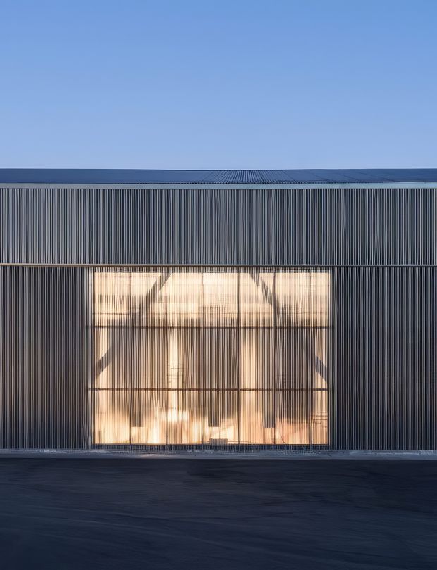 A stark, symmetrical exterior view of a contemporary building facade clad in closely spaced dark vertical metal fins. A large warm-glowing glazed opening reveals an illuminated interior and structural elements behind the screen. The building has a flat roofline, a restrained monochromatic palette, and a dark paved foreground beneath a clear blue sky.