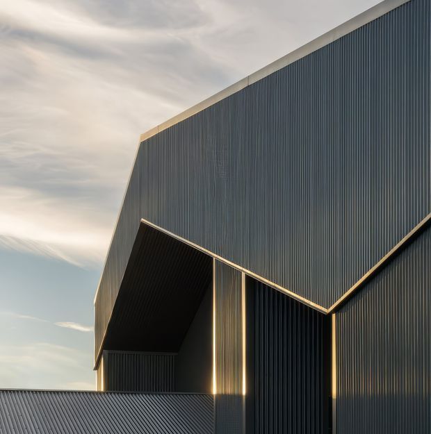 Close-up exterior architectural view of a contemporary building with bold angular geometry, deep recessed openings, and vertically ribbed dark metal cladding. Warm sunlight highlights the sharp roof and facade junctions against a softly clouded sky.