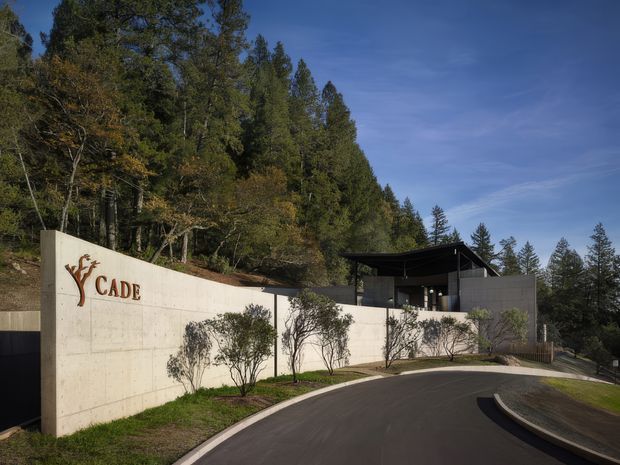 Exterior view of a contemporary facility set into a wooded, sloping site. A curved paved driveway leads past a tall smooth concrete retaining wall with the prominent CADE logo, toward a dark, flat-roofed pavilion-like building partially screened by the forest.
