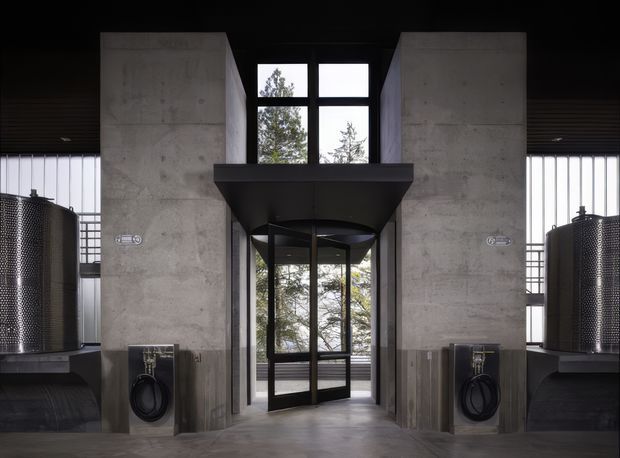 A dramatic industrial-style interior entry space framed by two tall raw concrete walls, with a black-framed glazed doorway and upper windows beneath a dark projecting canopy. Large cylindrical metallic tanks or equipment sit on both sides, alongside exposed industrial fixtures and dark ribbed ceiling and wall elements. Trees are visible through the glazing, but the focus is the architectural entrance and industrial fit-out.