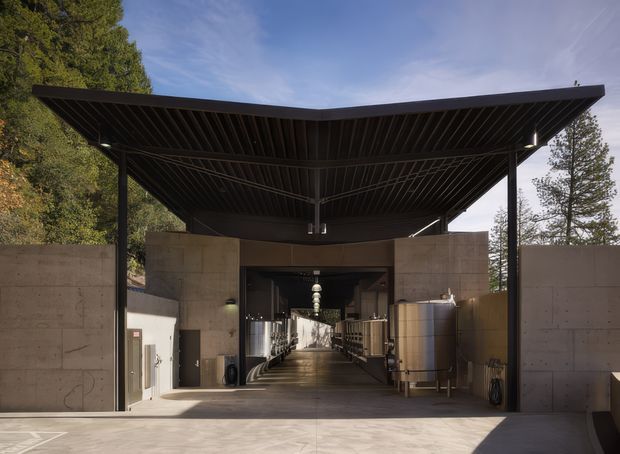 A striking modern architectural entrance or covered forecourt framed by exposed concrete walls and a large cantilevered dark metal roof canopy. The open central passage leads into a long industrial-looking interior with equipment and illuminated fixtures, while mature trees surround the structure. The image emphasizes strong symmetry, exposed structural elements, and a refined material palette of concrete, dark metal, and natural surroundings.