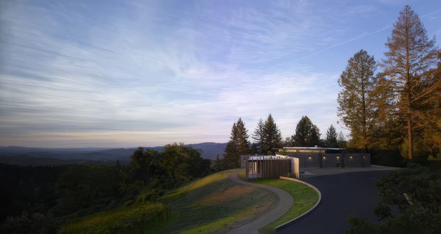 A dusk exterior view of a modern, low-profile residence set on a heavily wooded, elevated hillside. A curved driveway and walkway lead to a dark, minimalist building with a long horizontal roofline and vertical timber screening, while tall trees and expansive distant landscape surround the property.