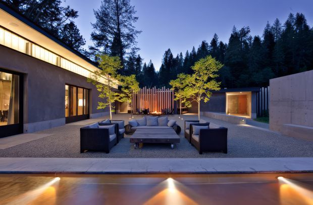 A twilight view of a contemporary courtyard and outdoor entertaining area, with a low flat-roofed building enclosing the space. The gravel-paved courtyard contains a large outdoor lounge arrangement with sofas, armchairs and a central coffee table, while a linear fire feature sits against a vertical timber screen. Warm architectural lighting highlights the building edges and a shallow reflecting pool or water feature occupies the foreground. Dense trees form the backdrop.