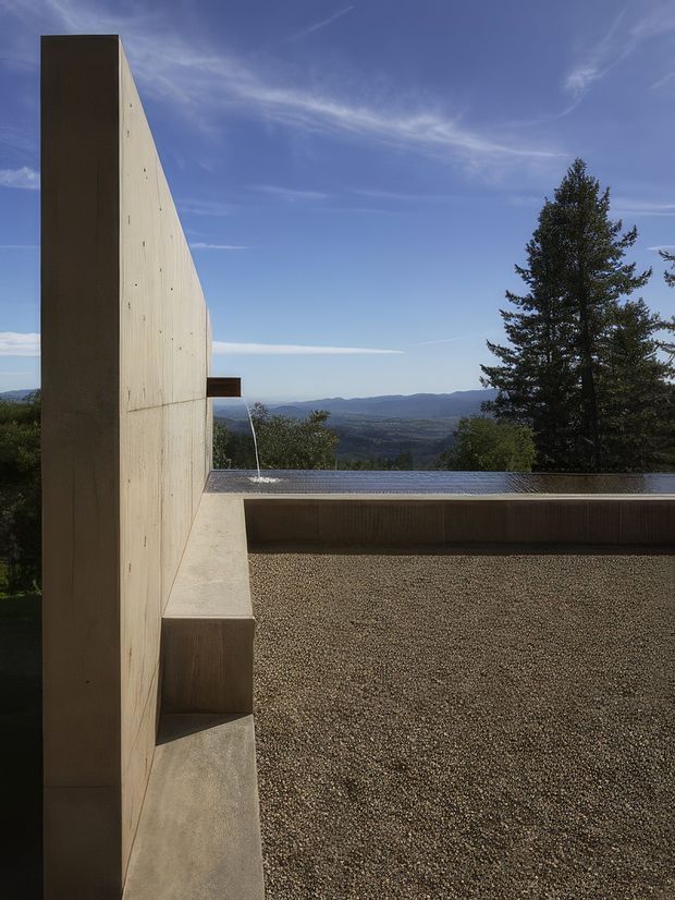 Exterior architectural view of a minimalist concrete structure on a hillside, featuring a tall blank concrete wall, flat roofline, gravel-covered terrace, narrow reflecting or swimming pool edge, and a distant mountainous landscape under a clear blue sky.