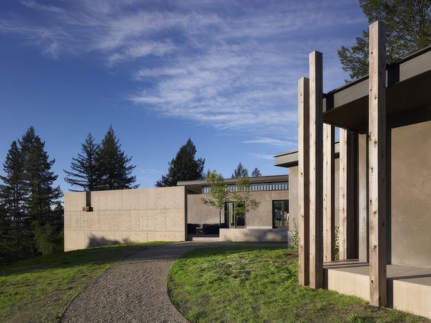 Exterior architectural photograph of a contemporary, low-profile house set within a landscaped, wooded site. The composition shows a gravel path leading toward a concrete and plaster building, with projecting flat roof forms, extensive glazing, and a prominent foreground screen of tall natural timber posts. Mature trees and lawn surround the structure under a clear blue sky.