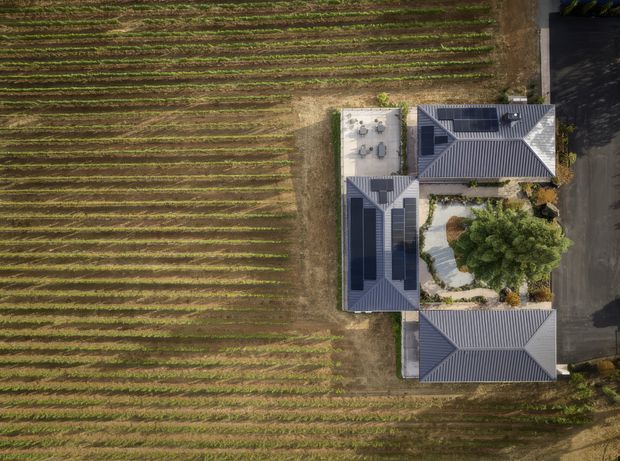 Aerial, top-down view of a large property beside an expansive cultivated field. The building complex has several connected wings with dark grey pitched, hipped-style roofing, a central landscaped courtyard with mature trees and curved pale pathways, and a small paved outdoor terrace. A driveway or road runs along the right edge.
