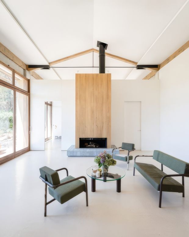 Bright, minimalist living room in a high-ceilinged pavilion-like interior. A central timber-clad fireplace chimney rises to the exposed pitched roof structure, with a black stove flue and dark fireplace opening. Large timber-framed glazing lines the left side, while a green upholstered sofa and matching armchairs surround a round glass coffee table. The palette is predominantly white with natural timber and muted green accents.
