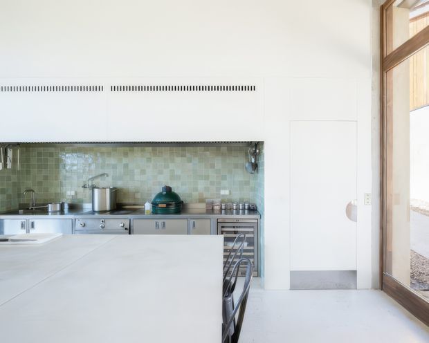 Bright, minimalist kitchen with a large pale island, stainless-steel cabinetry and appliances, and a muted green mosaic-tile splashback. Full-height timber-framed glazing opens the room to the outdoors, while white walls and integrated ventilation maintain a clean, contemporary appearance.