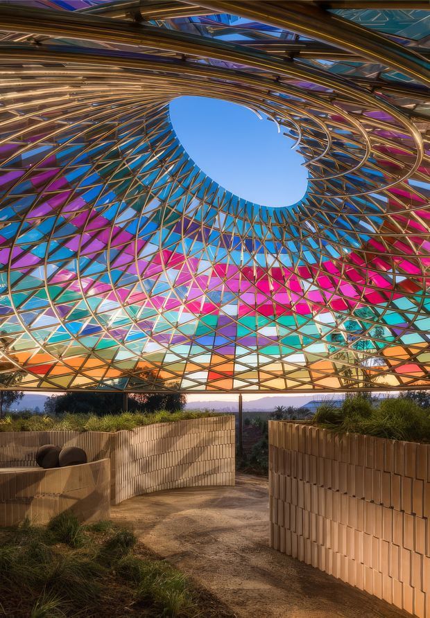 A dramatic outdoor architectural pavilion or walkway viewed beneath a large sculptural canopy. The canopy consists of a dense curved lattice of slender timber-like structural members with vivid translucent stained-glass or coloured panels in blue, magenta, purple, orange, and turquoise, forming an oval opening to the sky. Curved tan masonry or terracotta-toned screen walls line a sandy pathway, with planted grasses and distant landscape visible beyond. The image has a highly artistic, immersive, and organic character.