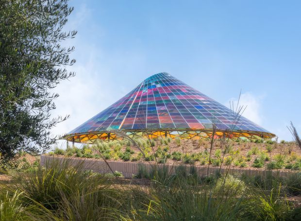 Exterior view of a distinctive conical, pyramid-like glazed structure with a colorful grid of blue, purple, pink, and red glass panels. The building has a broad projecting canopy supported by exposed steel trusses, set within landscaped native-style planting under a clear sky.