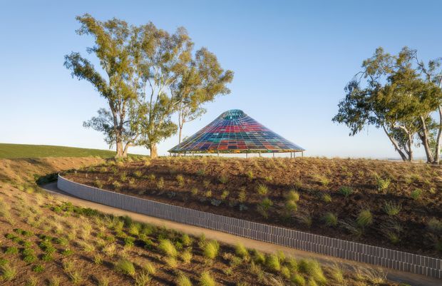A low, pyramid-shaped pavilion with a colourful translucent or glazed geometric roof sits within a landscaped sloping site. The structure is framed by mature trees and surrounded by planted earth banks, with a curving ribbed retaining wall or walkway edge in the foreground. The image is a bright exterior architectural landscape photograph under a clear sky.