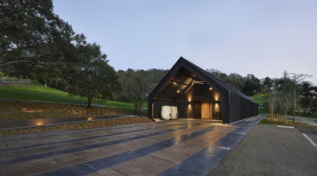 A dark, contemporary gable-roofed pavilion or barn-like building set within a landscaped rural site. The facade is clad in vertical charcoal material, with a large open gabled entry revealing warm timber elements, pendant lighting, and two white sculptural vessels. Broad wet stone or concrete paving leads toward the entrance, while mature trees, lawns, planted terraces, and subtle landscape lighting surround the building.