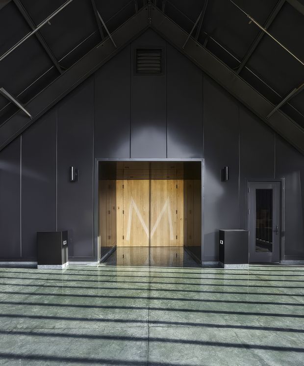 A dramatic, symmetrical interior entrance or lobby with a tall dark gabled ceiling and exposed structural framing. The charcoal wall and ceiling panels surround a large central opening with warm timber double doors and matching timber cabinetry beyond. A polished dark floor reflects the doorway, while strong linear shadows fall across the broad foreground floor. Minimal black wall lights, low dark joinery, and a side access door reinforce the industrial, refined character.