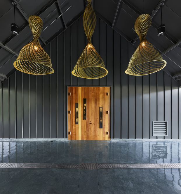 A dramatic, symmetrical entryway with a dark vertically ribbed metal wall and steeply pitched exposed roof structure. Three sculptural gold wire pendant lights hang above a warm timber double door. The polished dark floor reflects the lighting and facade, creating a luxurious, high-contrast atmosphere.