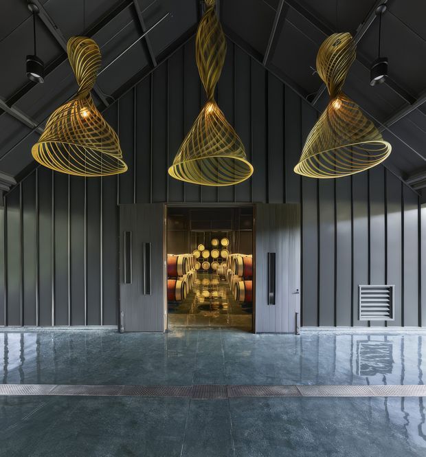 A dramatic dark industrial-style interior or entrance hall with vertically ribbed charcoal metal wall cladding, exposed structural roof framing, polished concrete flooring, and a central glazed doorway revealing a warmly lit interior with wine barrels. Three large sculptural gold wire pendant lights dominate the ceiling, creating a strong contrast against the black architectural backdrop.