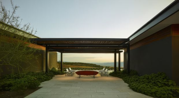 A modern courtyard terrace framed by low dark-clad wings and a flat-roofed, open steel pergola. Pale large-format paving leads to an outdoor seating area with a circular fire feature, while dense planting borders both sides. The open view beyond overlooks rolling countryside and distant hills.